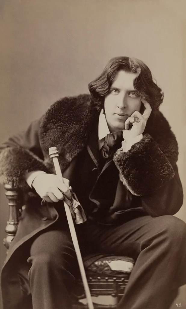 Photograph of Oscar Wilde (1882) in sepia tones, sitting in a chair with a walking cane and a fur-collared coat.