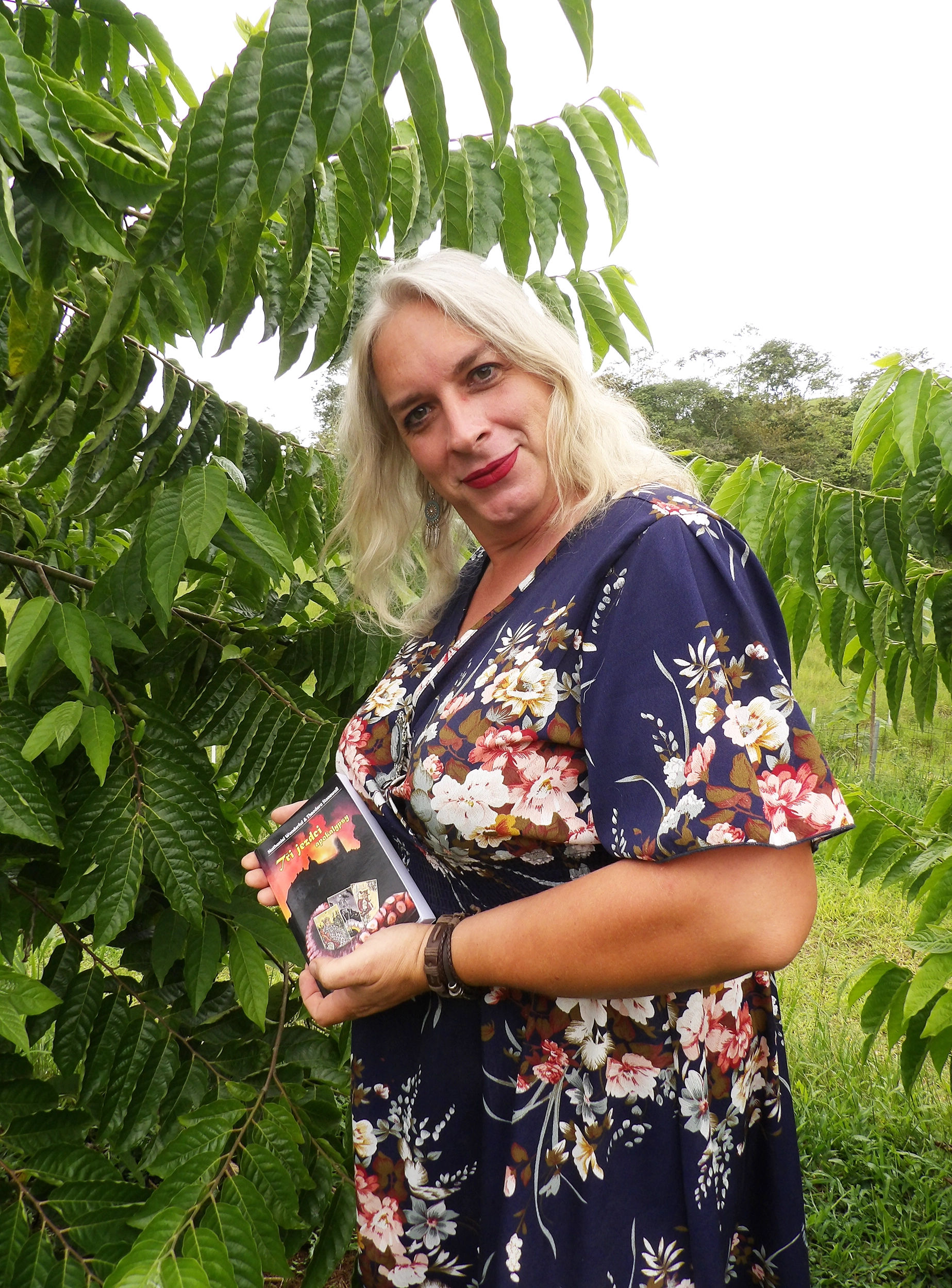 A trans woman with blonde hair in a dark blue floral dress, holding a book.